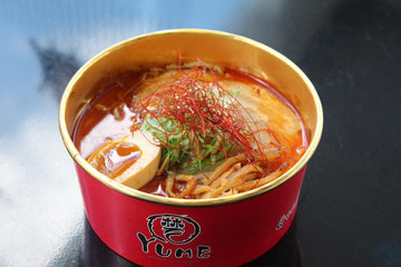 Bowl of ramen with spicy broth, tofu, vegetables, and noodles in a red Yume container